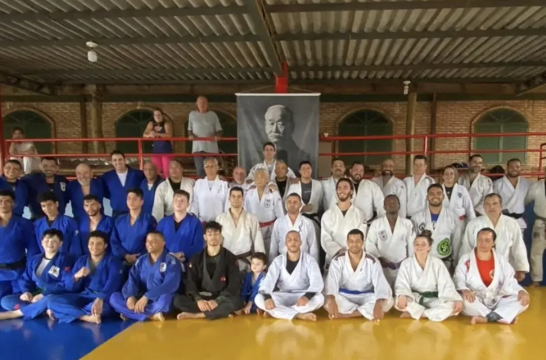 Cerca de quarenta judocas de quimono branco, azul e preto, sentados e em pé no tatame, sob um retrato de Jigoro Kano.