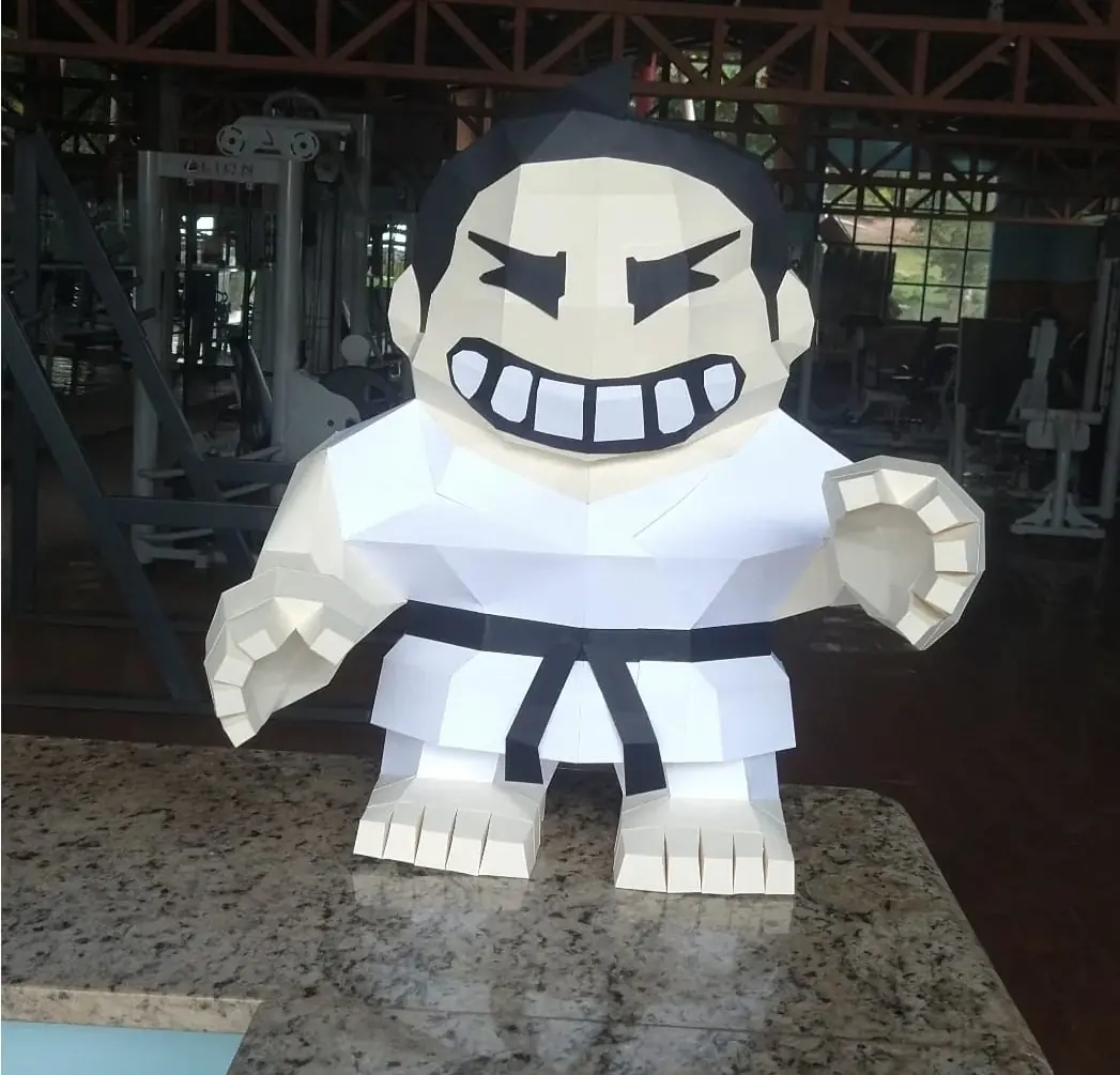 Escultura grande do mascote da academia — o judoca de quimono branco e faixa preta que aparece no logo — sobre uma bancada, com aparelhos ao fundo.
